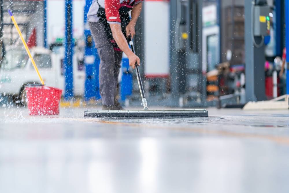 service staff man using a mop to remove water in the uniform cleaning the protective clothing of the new epoxy floor in an empty warehouse or car service center